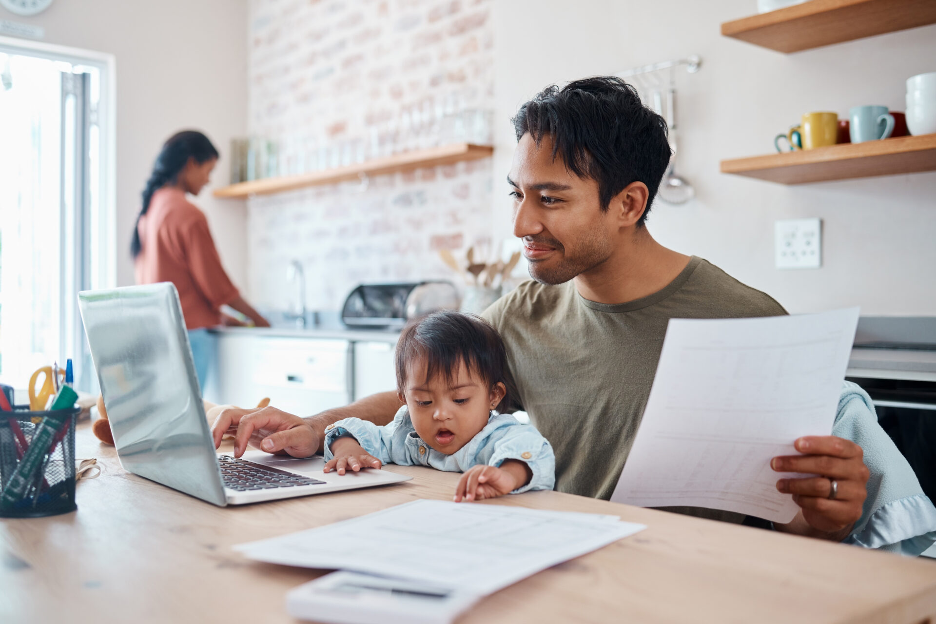 Father and young child sit at a bright kitchen table with a laptop and papers, while a woman washes dishes in the background.